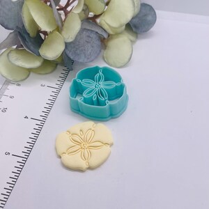 May include: A light blue plastic sand dollar cookie cutter next to a baked sand dollar cookie. The cutter is approximately 2 inches in diameter. The cookie is a light yellow color and has a detailed floral design.
