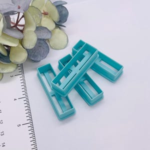 May include: A collection of five rectangular, light blue cookie cutters. Each cutter has a hollow center with a thin bar running across the middle. The cutters are arranged on a white surface, with a ruler and artificial greenery in the background.