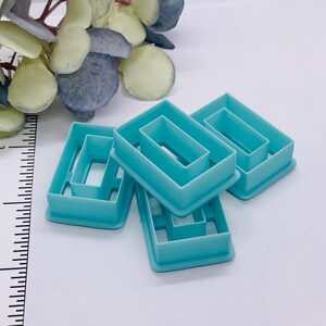 May include: Four teal-colored plastic cookie cutters in a rectangular shape with a smaller rectangle cut out of the center. The cutters are arranged on a white surface with a ruler and some artificial greenery in the background.