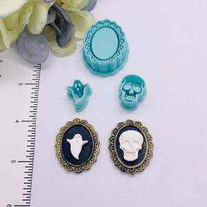 Op de afbeelding: Een verzameling Halloween-thema knutselspullen. Bevat een turquoise ovale mal, spook- en schedelvormige koekjessnijders en twee ovaalvormige broches met spook- en schedelontwerpen. De broches hebben een zwarte achtergrond en goudkleurige frames.