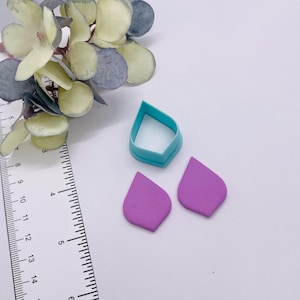 May include: A teal-colored polymer clay cutter and two purple clay teardrop shapes. A ruler with inch measurements is next to the clay pieces. Decorative flowers are in the background.