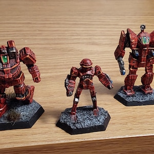 May include: Three miniature red and black mechs with detailed armour and weaponry. The mechs are standing on hexagonal black bases with textured surfaces. The mechs are painted with a weathered effect, giving them a battle-worn appearance. The mechs vary in size and design.