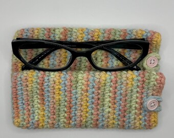 May include: A crocheted glasses case with pastel stripes in pink, yellow, blue, and green. The case has two pink buttons and a pair of black eyeglasses are inside.