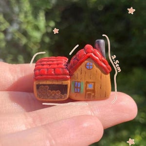 Può includere: Una casa in miniatura fatta a mano con tetto rosso e camino. La casa è dipinta in tonalità marroni e beige, con dettagli di finestre blu. La casa è alta circa 2,5 cm.
