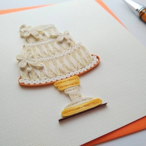May include: A handmade, three-tiered paper quilled cake with floral accents. The cake is cream-colored with orange and yellow details, set on a white card with an orange border. A pen rests in the background. This unique paper art piece is perfect for special occasions.