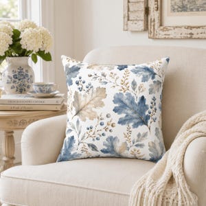 Könnte beinhalten: Ein quadratisches Zierkissen mit einem botanischen Muster auf weißem Hintergrund. Das Design zeigt Eichenblätter im Aquarellstil in Blau- und Beigetönen, durchsetzt mit kleinen blauen Beerenclustern und zarten beigen Zweigen.
