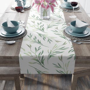Könnte beinhalten: Weißer Tischläufer mit einem Aquarelldesign aus grünen Blättern. Der Läufer liegt auf einem Holztisch mit blauen Gedecken und Weingläsern. Eine Vase mit rosa Lilien steht in der Mitte.
