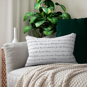 Peut inclure: Un coussin rectangulaire doté d'une housse en tissu blanc imprimée de texte en cursive noire reprenant le préambule de la Constitution des États-Unis. Le texte inclut des phrases telles que We the People of the United States, in Order to form a more perfect Union.