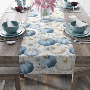 Puede incluir: Un mantel blanco con un estampado repetido de calabazas azules y blancas y detalles florales. La mesa está puesta con cuencos, platos y copas de vino de color turquesa. Un jarrón con lirios rosas está en el centro.