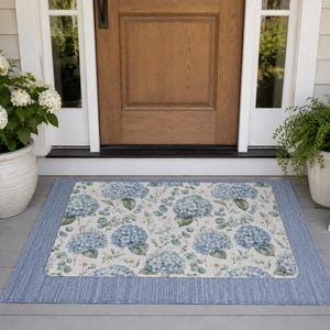 Puede incluir: Un felpudo rectangular con un panel central de estampado floral de hortensias azules y blancas, rodeado por un borde tejido de color azul sólido. El tapete está colocado en un porche gris frente a una puerta de madera con marco blanco.