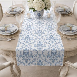 May include: A white table runner featuring a repeating blue damask pattern centered on a wooden dining table. The table is set with white plates, blue and white patterned bowls, folded napkins with gold rings, glassware, and a floral centerpiece.