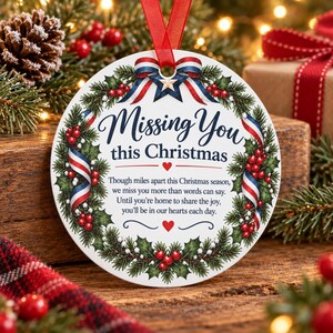 Può includere: Una decorazione natalizia rotonda in ceramica bianca con una ghirlanda di agrifoglio verde, bacche rosse e un nastro rosso, bianco e blu. Il testo recita: Missing You this Christmas, seguito da una poesia sulla mancanza e due cuori rossi.