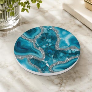 Op de afbeelding: Een ronde onderzetter met een levendig, op agaat geïnspireerd patroon in turquoise en blauwe tinten, geaccentueerd door textuurrijke zilveren glitterlijnen die de steenachtige secties op het oppervlak van de onderzetter verdelen.