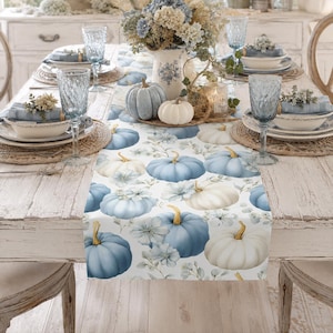 May include: A white table runner featuring a repeating pattern of light blue and cream-colored pumpkins interspersed with small floral accents. The runner is laid across a wooden dining table set with blue glassware, white plates, and pumpkins.