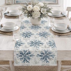 Op de afbeelding: Een witte tafelloper met een herhalend blauw aquarel sneeuwvlokpatroon op een lichthouten eettafel. De tafel is gedekt met witte keramische kommen, gevlochten placemats, heldere glazen en een centraal wit bloemstuk in een vaas.