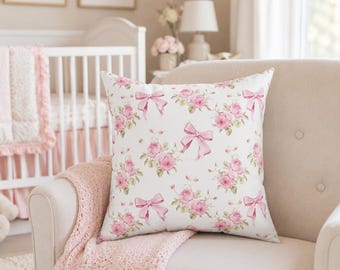 Puede incluir: Un cojín cuadrado con fondo blanco y un patrón repetitivo de rosas de color rosa suave y lazos de color rosa claro. El diseño está impreso en la parte frontal del cojín, mostrando racimos de flores y detalles de cintas atadas.