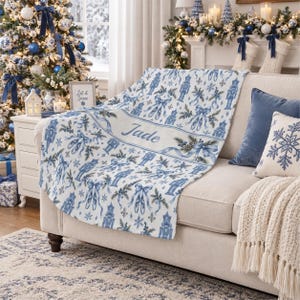 May include: A white throw blanket featuring a repeating blue pattern of nutcrackers, ballet slippers, bows, and pine branches. The blanket includes a central horizontal band with the name Jade printed in a blue script font across the center.
