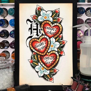 Op de afbeelding: Ingelijste tattoo-kunst met drie rode hartvormige ontwerpen, gouden randen en de zinnen "He wasn't shot", "didn't win" en "is on the list". Het ontwerp bevat witte bloemen en groene bladeren.