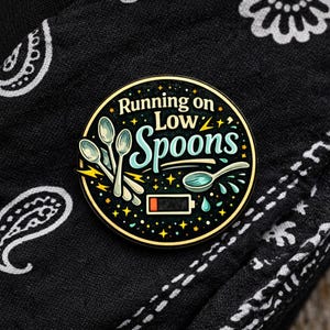 Puede incluir: Un pin esmaltado circular con fondo negro que presenta el texto Running on Low Spoons en blanco y azul claro. El dise&ntilde;o incluye ilustraciones de cuatro cucharas plateadas, rayos amarillos, peque&ntilde;as estrellas y un icono de bater&iacute;a baja.