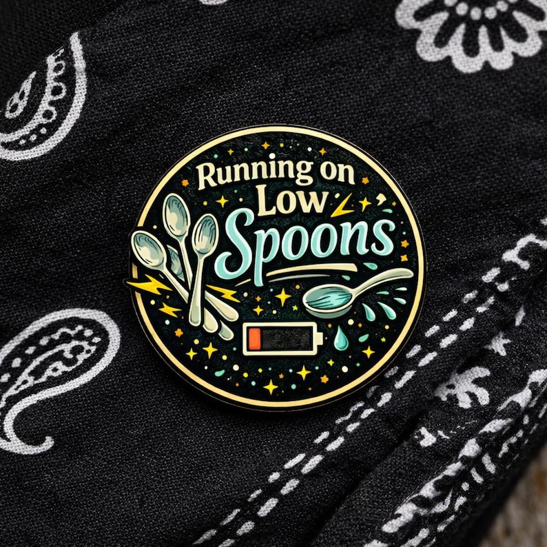 Puede incluir: Un pin esmaltado circular con fondo negro que presenta el texto Running on Low Spoons en blanco y azul claro. El dise&ntilde;o incluye ilustraciones de cuatro cucharas plateadas, rayos amarillos, peque&ntilde;as estrellas y un icono de bater&iacute;a baja.