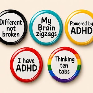 May include: Five circular pinback buttons with white centres and black text. The buttons feature the phrases: Different not broken, My Brain zigzags, Powered by ADHD, I have ADHD, and Thinking ten tabs. The borders are black, teal, yellow, red, and rainbow.