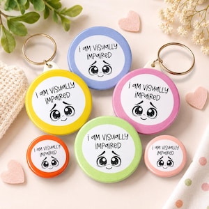Peut inclure: Un ensemble de six badges ronds et porte-clés de différentes couleurs, notamment jaune, bleu, rose, orange, vert et rose clair. Chaque objet porte l'inscription "i AM ViSUALLY iMPAiRED" au-dessus d'un visage de dessin animé aux grands yeux.