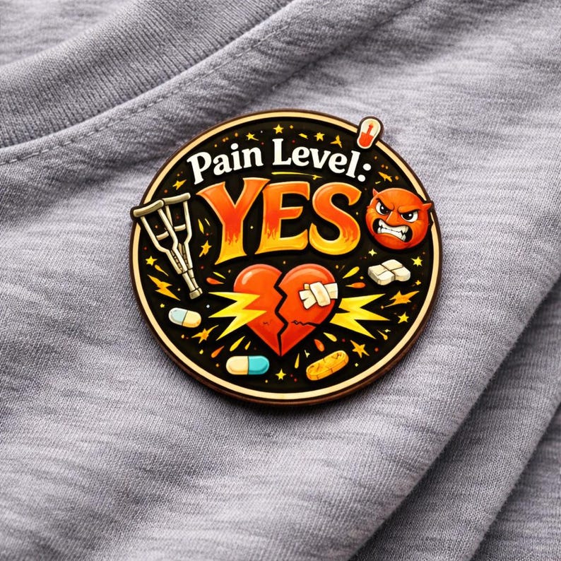 Puede incluir: Un pin esmaltado circular con fondo negro y el texto Pain Level: YES en naranja y blanco. El dise&ntilde;o incluye un par de muletas, un coraz&oacute;n roto con vendas, una cara roja enfadada y varias pastillas y c&aacute;psulas de colores variados.