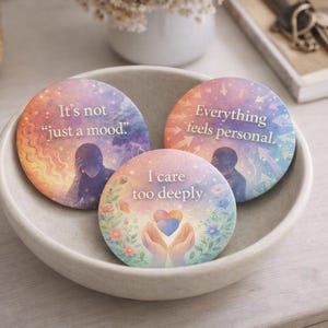 May include: Three round, colorful coasters with inspirational quotes in a white bowl. The coasters feature watercolor designs with phrases like "It's not just a mood," "Everything feels personal," and "I care too deeply."