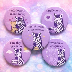 Puede incluir: Un conjunto de cinco botones redondos morados con ilustraciones de cebras. Cada botón tiene un mensaje diferente, incluyendo "Soft doesn't mean weak" y "I believe you". Los botones están decorados con corazones, estrellas y un calendario.
