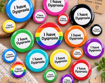 May include: A collection of round pin-back buttons in various colors including blue, pink, black, green, orange, yellow, purple, and red, featuring a rainbow striped button in the center. Each button displays the text I have Dyspraxia.