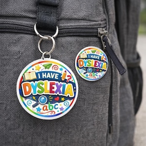 Puede incluir: Un llavero redondo y un pin con el texto "I HAVE DYSLEXIA" en letras coloridas. El llavero está sujeto a una mochila. El pin y el llavero tienen un fondo blanco con ilustraciones coloridas de un libro, un lápiz y una lupa.