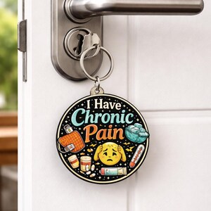 Puede incluir: Un llavero redondo con el texto I Have Chronic Pain en letras azules y naranjas. El dise&ntilde;o incluye ilustraciones de una bolsa de agua caliente, un cuenco de hielo, un term&oacute;metro, frascos de pastillas y una cara emoji amarilla sujet&aacute;ndose la cabeza.