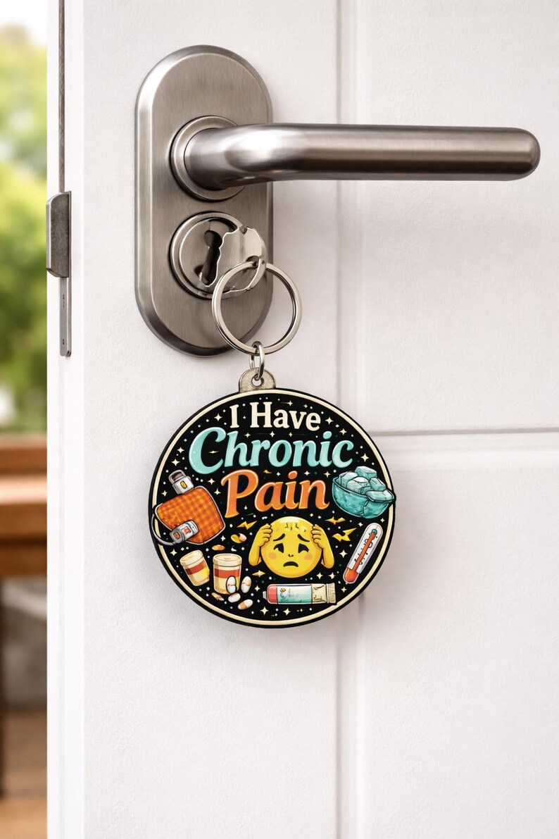 Puede incluir: Un llavero redondo con el texto I Have Chronic Pain en letras azules y naranjas. El dise&ntilde;o incluye ilustraciones de una bolsa de agua caliente, un cuenco de hielo, un term&oacute;metro, frascos de pastillas y una cara emoji amarilla sujet&aacute;ndose la cabeza.