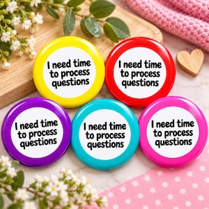 May include: Five circular pin-back buttons in yellow, red, purple, teal, and pink. Each button features a white center with the black text I need time to process questions. The buttons are arranged in a group on a light-colored surface.