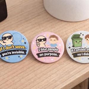 May include: Three circular badges with funny slogans and cartoon illustrations. One is blue and says "If I don't wave, you're invisible." Another is pink with "I blur people on purpose." The third is white with "If I smiled at no I didn't."