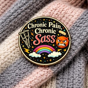 Puede incluir: Un pin esmaltado redondo con fondo negro que presenta el texto "Chronic Pain, Chronic Sass" en blanco y rosa. El dise&ntilde;o incluye dos muletas plateadas, una tirita beige, una peque&ntilde;a cara naranja enfadada, un arco&iacute;ris y estrellas amarillas.