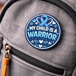 Puede incluir: Un pin redondo azul y blanco con el texto "MY CHILD IS A WARRIOR". El diseño incluye una cinta azul, corazones y elementos decorativos. Está sujeto a una mochila gris.