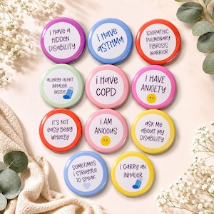 May include: A collection of round, colorful pinback buttons with various phrases related to health conditions. The buttons feature text like "I Have Asthma," "I Have Anxiety," and "Ask Me About My Disability." The buttons are arranged on a soft, neutral-colored surface.