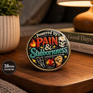 Puede incluir: Un pin redondo de 38 mm de di&aacute;metro sobre fondo negro con el texto Powered by PAIN & Stubbornness. El dise&ntilde;o incluye una llama, una calavera con una tirita, dos muletas, un coraz&oacute;n roto y un pu&ntilde;o cerrado en amarillo, rojo y azul verdoso.