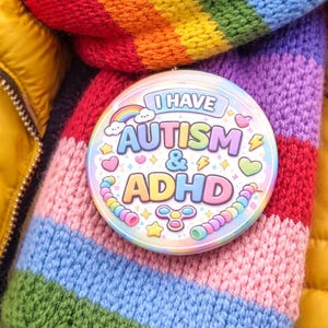 May include: A circular badge with the words "I HAVE AUTISM & ADHD" sits on a rainbow-striped scarf. The badge includes a rainbow, hearts, stars, and a fidget spinner. The scarf is made of knitted material.
