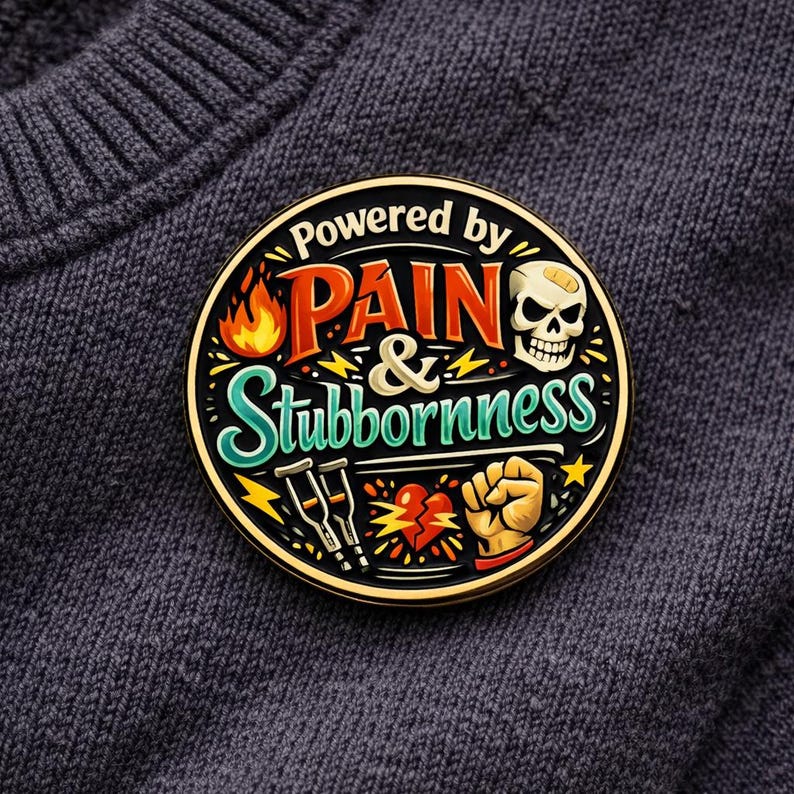 Puede incluir: Un pin esmaltado circular con el texto Powered by PAIN & Stubbornness en naranja, blanco y turquesa. El dise&ntilde;o incluye una calavera con una tirita, una llama, dos muletas, un coraz&oacute;n roto y un pu&ntilde;o cerrado sobre un fondo negro.