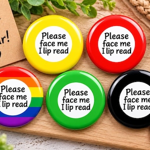 Puede incluir: Cinco chapas redondas en amarillo, rojo, arcoíris, verde y negro. Cada chapa presenta un círculo blanco central con el texto negro 'Please face me I lip read'. Las chapas están dispuestas sobre una superficie de madera con etiquetas decorativas.
