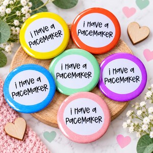 May include: A set of six circular pin-back buttons in yellow, orange, light green, blue, purple, and pink. Each button features a white center with the black text "i Have a paceMaker" printed in a casual, handwritten-style font.