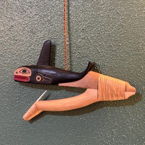 Può includere: Scultura in legno intagliato di un'orca nera e naturale. L'orca ha una pinna dorsale nera, dettagli rossi e dorati. La scultura è sospesa da un cordoncino marrone, che mostra l'artigianato tradizionale.