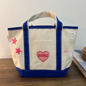 May include: Cream-colored tote bag with blue trim and handles. Features a front pocket and a heart-shaped patch with the word "BOOKS." Decorated with pink star and flower appliqués, and a white letter "P."