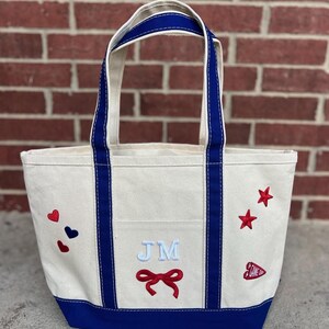 May include: A canvas tote bag with a cream-colored body, blue trim, and handles. The bag features embroidered red hearts and stars, the initials "JM" in light blue, and a red bow. A small patch reads "GAME ON".