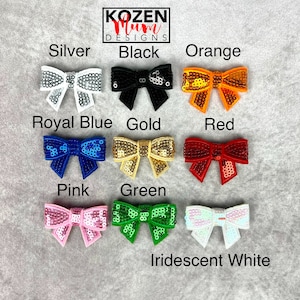 May include: A collection of sequined bow embellishments in various colors, including silver, black, orange, royal blue, gold, red, pink, green, and iridescent white. The bows are arranged on a gray surface, with each color labeled.