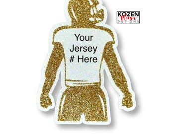May include: A gold glitter football player silhouette sticker featuring a white center panel with the black text Your Jersey # Here. The design includes a helmet, jersey, and pants, with a small logo in the corner reading KOZEN Mum DESIGNS.