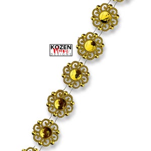 May include: A decorative gold trim featuring ornate, flower-shaped embellishments with a central, amber-colored gem. The trim is strung on a thin, silver-colored wire. The text "KOZEN MUM DESIGNS" is visible.