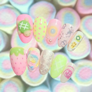 May include: A set of ten artificial fingernails featuring diverse designs including a pink nail with a green turtle, a yellow nail with pink dots, a pink nail with a central circle, a white nail with floral patterns, and a strawberry-themed nail.